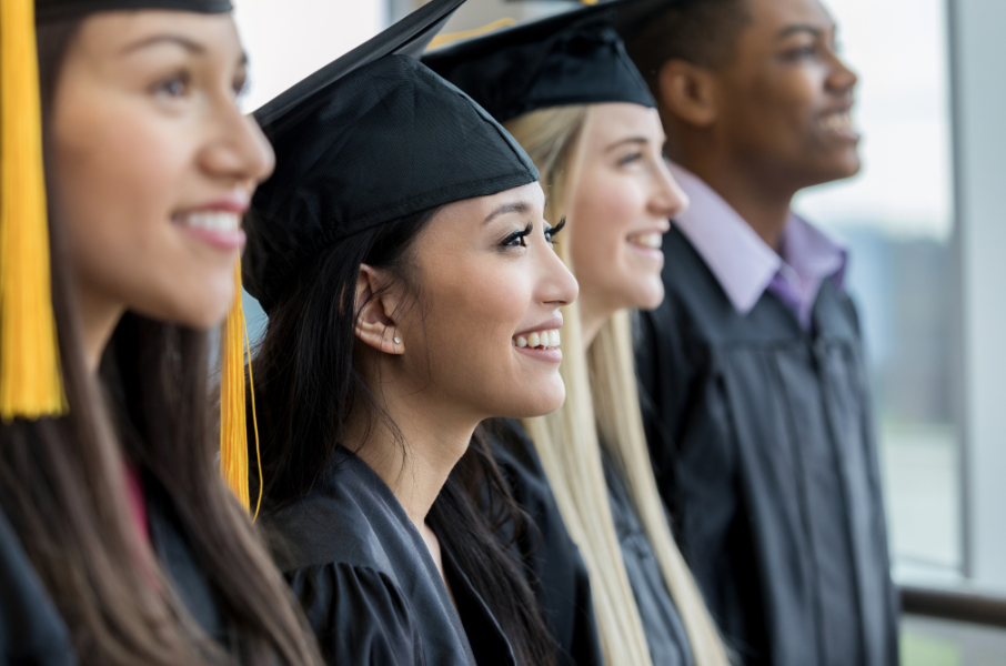 Four students sit excitedly at graduation ceremony dressed in caps and gowns